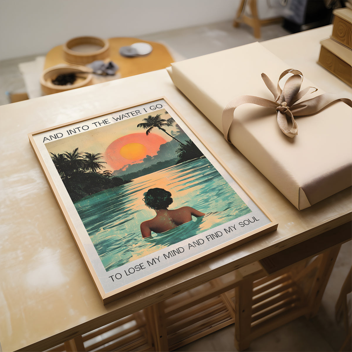 Poster of a woman in water with palm trees and sunset, placed on a table next to a wrapped gift.