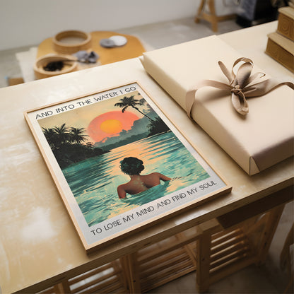 Poster of a woman in water with palm trees and sunset, placed on a table next to a wrapped gift.