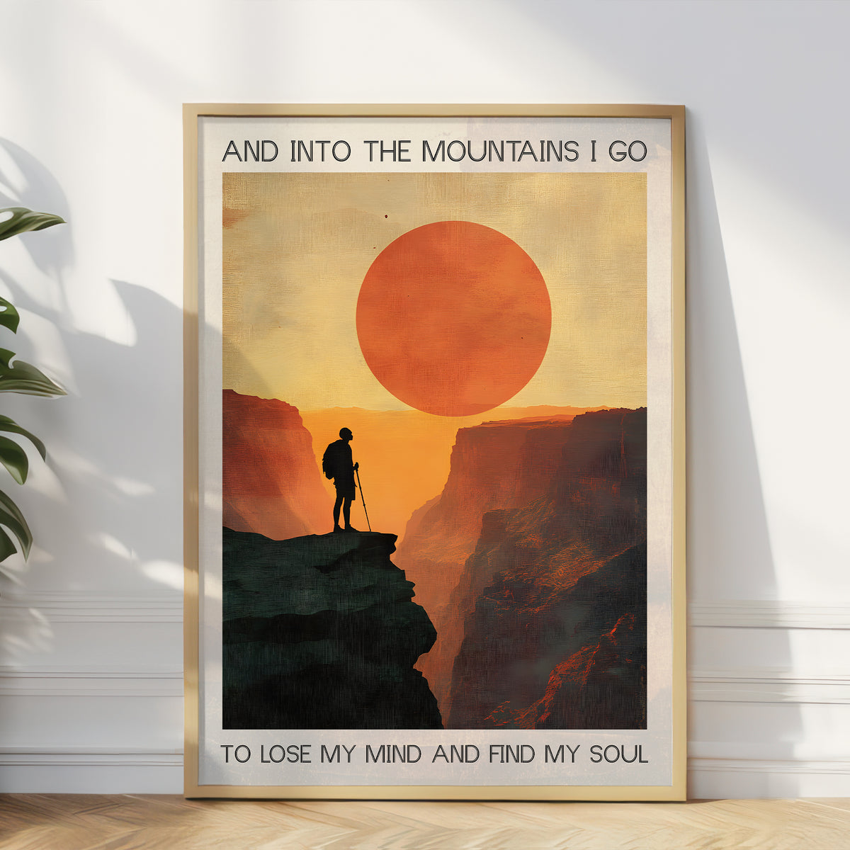 Framed poster of a silhouette standing on a mountain with a large orange sun, text 'And into the mountains I go to lose my mind and find my soul', on a white wall.
