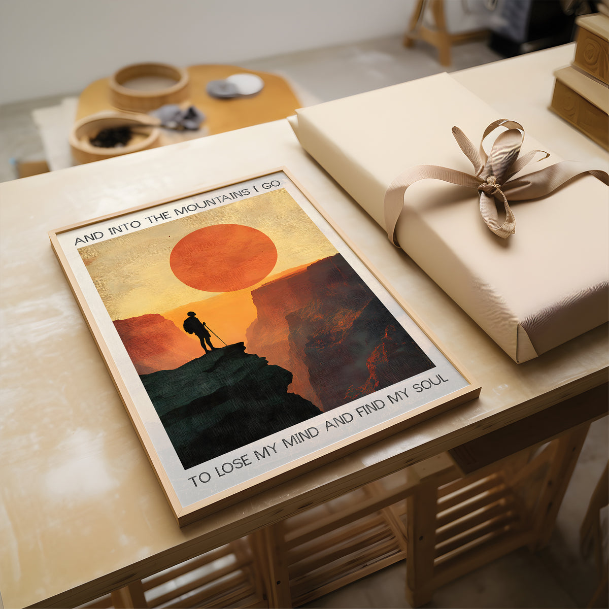 Poster of a mountain scene with a person on a rock, placed next to a wrapped gift on a wooden table.
